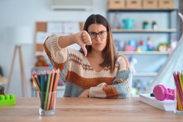 Young beautiful teacher woman wearing sweater and glasses sitting on desk at kindergarten looking...