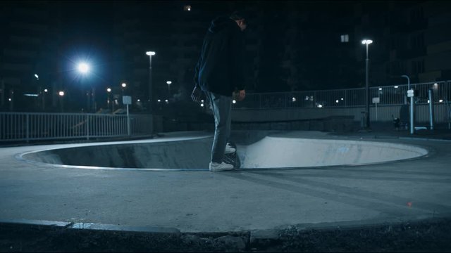 A Young Guy Comes To The Pool With A Skateboard And Prepares To Jump Into The Pit. Skateboarder Rides A Skateboard At Night On The Street, Lights Are On. Teenager On A Skateboard. Rear View, 4K