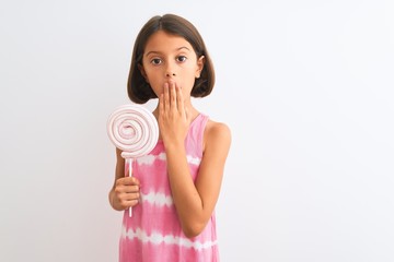 Beautiful child girl eating sweet lollipop standing over isolated white background cover mouth with hand shocked with shame for mistake, expression of fear, scared in silence, secret concept