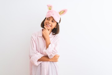 Beautiful child girl wearing sleep mask and pajama standing over isolated white background looking confident at the camera smiling with crossed arms and hand raised on chin. Thinking positive.