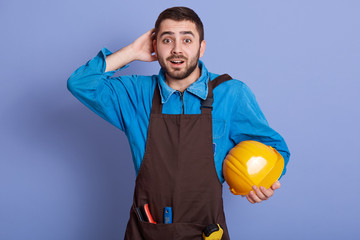 Portrait of surprised shocked construction worker putting hand at back of his head, looking directly at camera with astonishment, holding yellow helmet, having special equipment. Job concept.