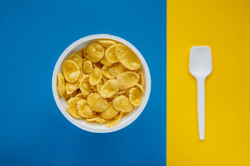 Cornflakes in white bowl with spoon on blue and yellow background. Top view. Copy, empty space for text