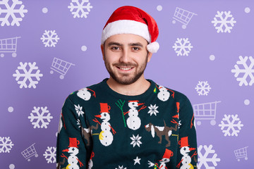 Indoor studio shot of handsome bearded man wearing red santa claus hat and new year sweater with snowmen, having peaceful facial expression, looking directly at camera, having snowdrops at background.