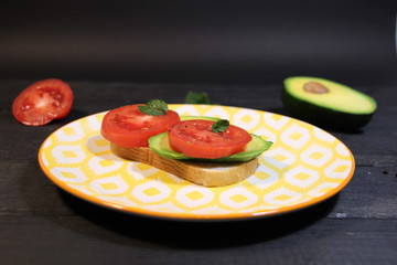 Open vegetarian sandwich with slices of tomato, avocado, mint, bread, pepper, salt, oil on bright plate on black wooden background