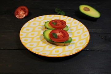 Open vegetarian sandwich with tomato, avocado, mint, bread, peppert, salt, oil on bright plate on black wooden background