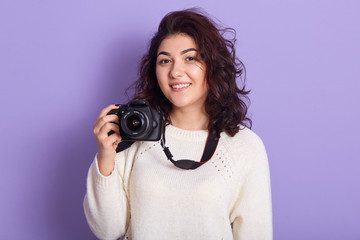 Picture of charming magnetic young female with curly black hair, holding photo camera, taking photos, smiling sincerely, wearing white sweater, spending time alone, getting pleasure. Hobby concept.