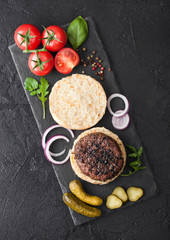 Fresh grilled minced pepper beef burgers on stone chopping board with buns onion and tomatoes on black background. Salty pickles and basil