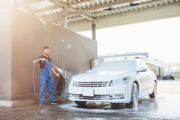 Professional bearded  washer in blue uniform washing luxury car with water gun on an open air car...