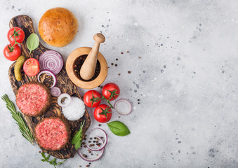 Fresh raw minced pepper beef burgers on vintage chopping board with buns onion and tomatoes on wooden background.Mortar with pestle with pickles and basil.