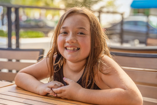A little girl smiles at the camera while sitting at a table outside on the cafe patio. A beautiful day to be outdoors enjoying the warm sunlight while on vacation.