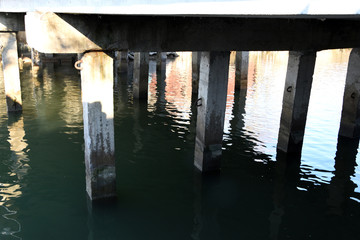 Concrete piles of a column, a reflection of the environment in the water, are visible in rows in the water. The sun's rays fall on the gray pillars.