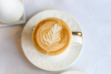 Cup of cappuccino with a beautiful pattern on the foam on the white plate and the table