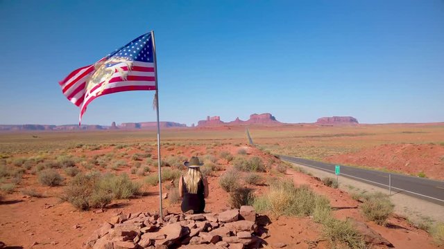 Young blonde girl sits near USA waving flag in Forrest Gump Point Road to Monument Valley, region of Colorado Plateau buttes Arizona&ndash;Utah border. Slow motion Gimbal panning cinematic ProRes 422