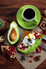 A healthy tasty breakfast with an avocado sandwich, poached egg, pomegranate seeds, soft cheese and coffee on a brown wooden board. Near pieces of pomegranate