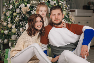 Beautiful young family sitting next to a nicely decorated Christmas tree. Happy family having fun with Christmas presents at home. Christmas at home by the fireplace and the Christmas tree. Portrait