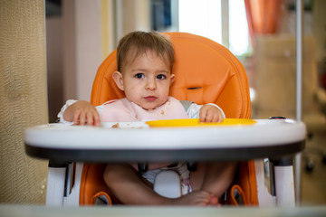 cute Caucasian baby girl sitting in an orange baby chair after eating with crumbs on her face, on the table crumbs and an orange plastic plate. close-up, front view, soft focus, blurred background