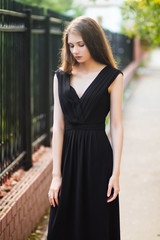 young, beautiful girl in black dress, outdoor photo on a background of steel fence, summer evening. Moddel with clean skin.