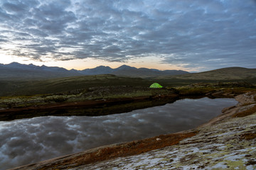 Green tent lighted up outdoors in the mountains by lake during blue hour. Blue clouds in the background, mountain scenery. Wilderness and explorer concept.