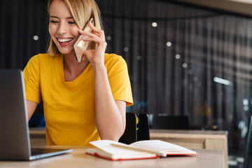 Photo of cheerful young woman using laptop and talking on cellphone