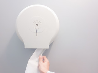 Hand-pulling the tissue roll out a white box mounted on a white wall in a public restroom.