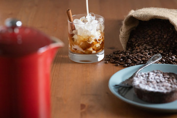Milk pouring into a glass with Cold brew coffee, crushed ice and a cinnamon stick surrounded by a bag of coffee beans, cake and a french press