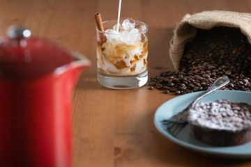 Milk pouring into a glass with Cold brew coffee, crushed ice and a cinnamon stick surrounded by a bag of coffee beans, cake and a french press
