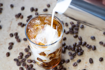 Milk pouring into a glass of iced coffee with beans in the blurry background