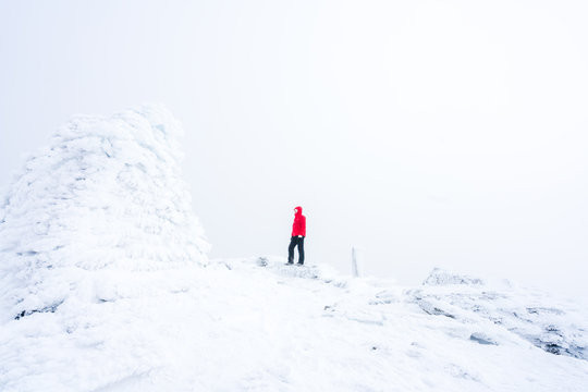 A man/hiker with red jacket and norwegian flag on his chest standing on a snowy mountain peak next to a frozen cairn. Trekking and adventure concept.