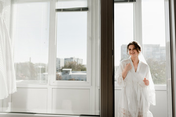 Portrait of young gorgeous bride on the balcony. Beautiful girl dresses and gathers for a wedding ceremony in apartments while waiting for the groom. Bride looking at her dress. Bride's morning