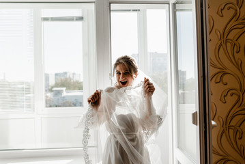 Portrait of young gorgeous bride on the balcony. Beautiful girl dresses and gathers for a wedding ceremony in apartments while waiting for the groom. Bride looking at her dress. Bride's morning