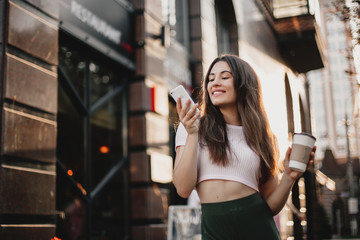 Smiling woman using phone and drink coffee on the street in summer day.