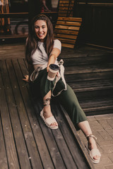 Happy smiling woman drink coffee sitting on wooden steps in street.