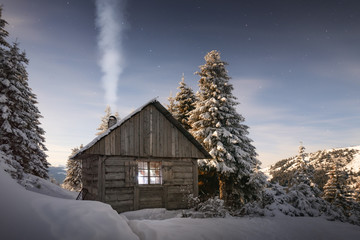Fantastic winter landscape with wooden house in snowy mountains. Smoke comes from the chimney of snow covered hut. Christmas holiday concept