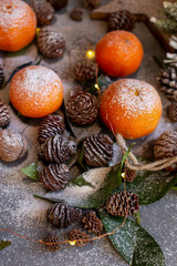 Orange tangerines on grey background in New Year's decor with brown pine cones and green leaves. Christmas decoration with mandarins. Delicious sweet clementine.