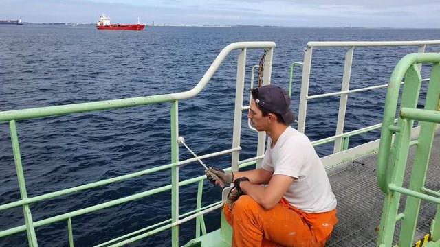 sailor in rubber gloves sits on haunches and paints railings white color against calm dark blue sea with red tanker