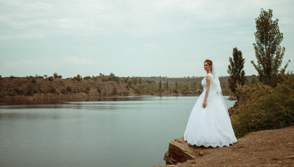 Fototapeta na wymiar A young smiling and beautiful bride in a white dress is standing against a background of rocks, cliffs and stones. Wedding portrait of a cute curly bride. Top vew. Space for text.