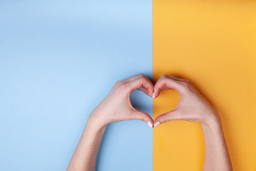 Beautiful female hands with perfect manicure make a heart on a blue-yellow background. Place for...