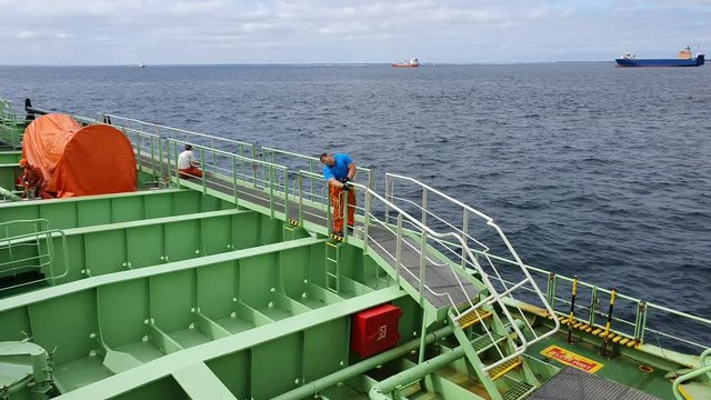 Onyx tanker sailor works on deck in orange coveralls and blue t-shirt against calm sea with distant vessels on raid