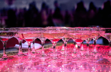 Many Champagne glasses at party are lined up ready to be served. Celebrate, cheer, cocktail in the background. Elegant catering, tiered alcohol drinks on glass table in luxury purple concept