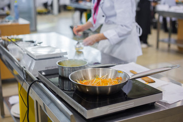 Process of cooking sliced carrots, cabbages and peppers in frying pan with oil on electric stove at cuisine of restaurant. Professional cooking, catering, cookery, gastronomy and food concept