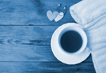 Cozy flatlay composition with white knitted scarf, cup of tea, two decorative heart on wooden desk. Christmas, Valentines day tredy color blue concept. Copyspace.