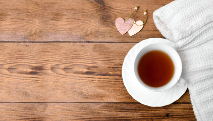 Cozy flatlay composition with white knitted scarf, cup of tea, two decorative heart on wooden desk. Christmas, Valentines day or hygge winter holiday concept. Copyspace.