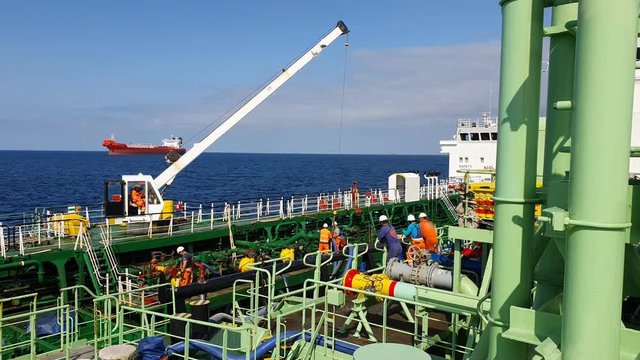 big oil tanker hoist helps ship workers to set bunker hoses on deck against dark blue sea under clear sky