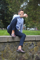 Obraz premium Portrait of a guy in a shirt sitting in a park in a European city. A man in the background of trees and buildings. Stock photos