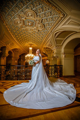 Luxury blonde bride with a white bouquet stands in a golden interior next to the chandelier