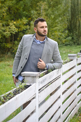 Portrait of a sexy man dressed in a suit. Fashionable guy in the autumn nature near a white wooden fence. Businessman relaxes. Skok photo for design © snb2087