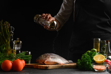 Chef cooks fresh fish on the background with ingredients, salts it before cooking, freezing in motion, advertising photo for seafood sale, cookbook for recipes