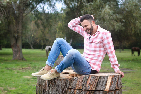 Young handsome guy Cowboy. farmer is sitting on a large stump on his ranch. Rural landscapes, countryside. Trees, field, farm. Stock photos for design.