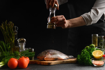 Chef cooks fresh fish on the background with ingredients, peppers it before cooking, freeze in motion, advertising photo for seafood sale, cookbook for recipes, healthy food