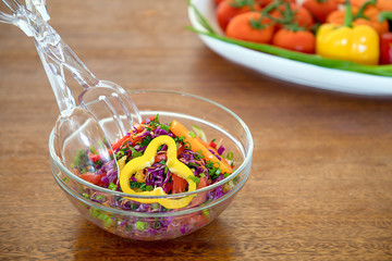A glass bowl with vegetable salad on wooden table, transparent serving tongs, a white dish with veggies on background